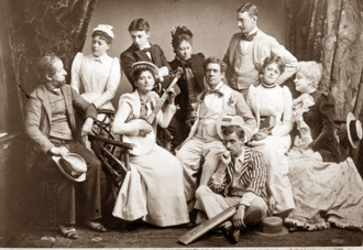 group of ten white people of both sexes and various ages in late Victorian costumes; four young people stand in the back row, five people of mixed ages, one of them holding a banjo, sit in the middle row, and a young man holding a cricket bat sits on the floor at their feet