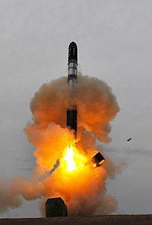 A rocket climbing out of a hole in the ground through an orange cloud of dust and smoke, with a metal ring falling away from the bottom of the rocket