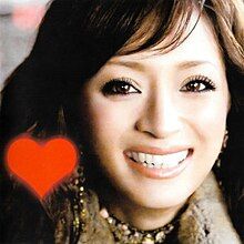 An up-close shot of Ayumi Hamasaki smiling and looking into the camera. On the lower left is a red heart.