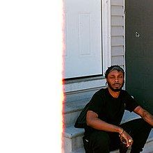 JPEGMAFIA sitting on a porch in front of a house with grey walls and a white door, he is holding a drink and his eyes are closed. Half of the photo is completely white due to a first of the (film) roll burn effect