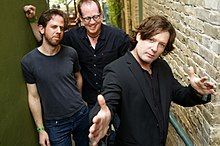 Three men wearing black shirts stand in a hallway.