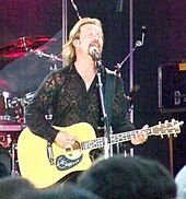 A man with long hair and a beard, wearing a black shirt, playing a guitar and singing into a microphone