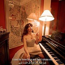 Artist Sarah Bahbah sits in front of a piano. She is wearing a formal strapless white dress, holding a lit cigarette. She has long, wavy brown hair and bangs. To her right is a lit lamp, and there is a large framed art piece behind her.