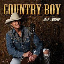 A photo of the artist wearing a denim jacket and white cowboy hat, sat on a stool in front of a brown background and holding an acoustic guitar.