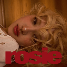 Close-up of Rosé lying down with blonde curly hair, gazing at the camera. The word ‘rosie’ is written in bold red text across the bottom of the image.