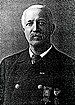 Head and shoulders of a white man with mustache wearing a double-breasted suit coat over a shirt and tie. Two medals are pinned to the coat's left breast.