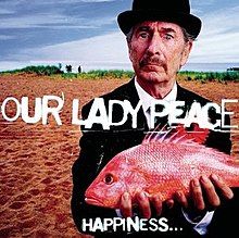 An old man standing on a sandy beach wearing a black suit and bowler derby stands holding an orange-pink fish in both hands. He stares at the audience with a puzzled look on his face.