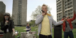 A young woman in a dark leather jacket, a man in a blue jumper, a man with bleached blonde hair in a yellow T-shirt and a man in a red leather jacket are performing on a grassy area outside a tower block estate during daytime. The man in the blue jumper is drumming at a drum kit; the other three are singing into microphones.