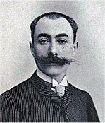 Head and shoulders photo of a youngish man with receding dark hair and large moustache, wearing striped suit