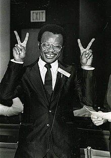 A black and white photograph of a man in a suit posing with two V signs