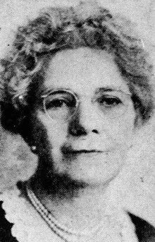 A woman with grey curls, wearing glasses, pearls, and a dark top with a white lace trimmed neckline