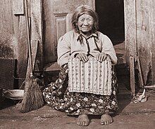 An elderly Clatsop woman sitting in front of a wooden door, wearing western style clothes. She is holding a traditional basket.