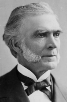 A black-and-white, head-and-shoulders photographic portrait of an elderly Caucasian gentleman with greying hair and white mutton chop sideburns. He is smartly dressed in a dark suit jacket and white shirt with a Gladstone collar and Kentucky-style bowtie.