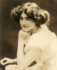 A young white woman with dark hair in a bouffant updo, seated with one hand on chin