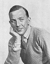 smiling, seated, clean-shaven young white man in tie and jumper, resting chin on hand